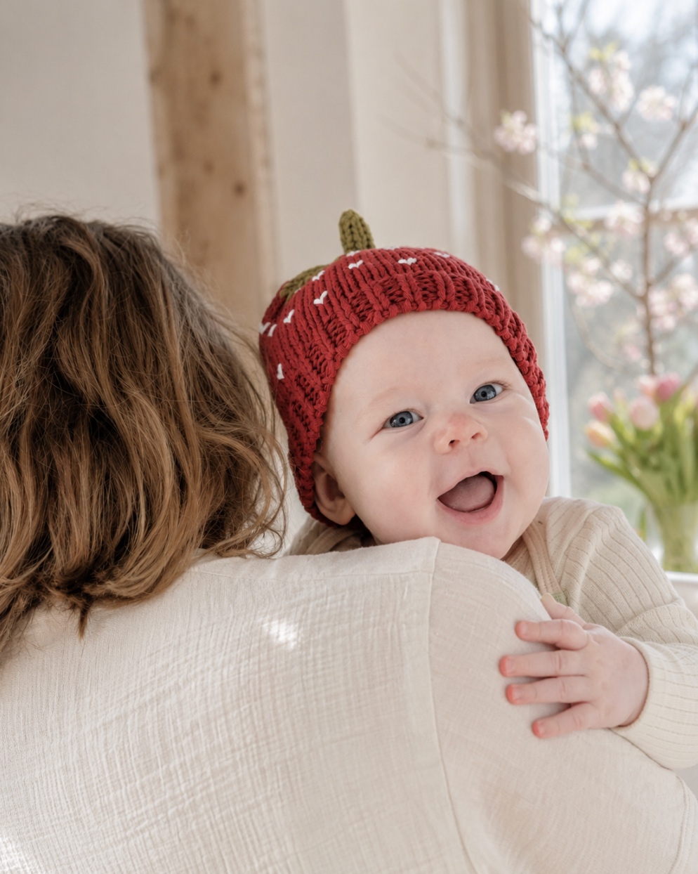 Smiling baby wearing the red Tiny & Wild Strawberry Beanie while being held